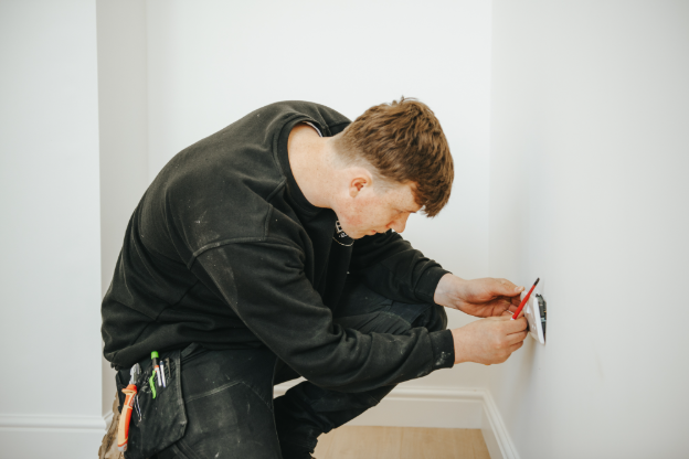 An electrician knelt on the floor, holding a screwdriver and working on a plug socket.